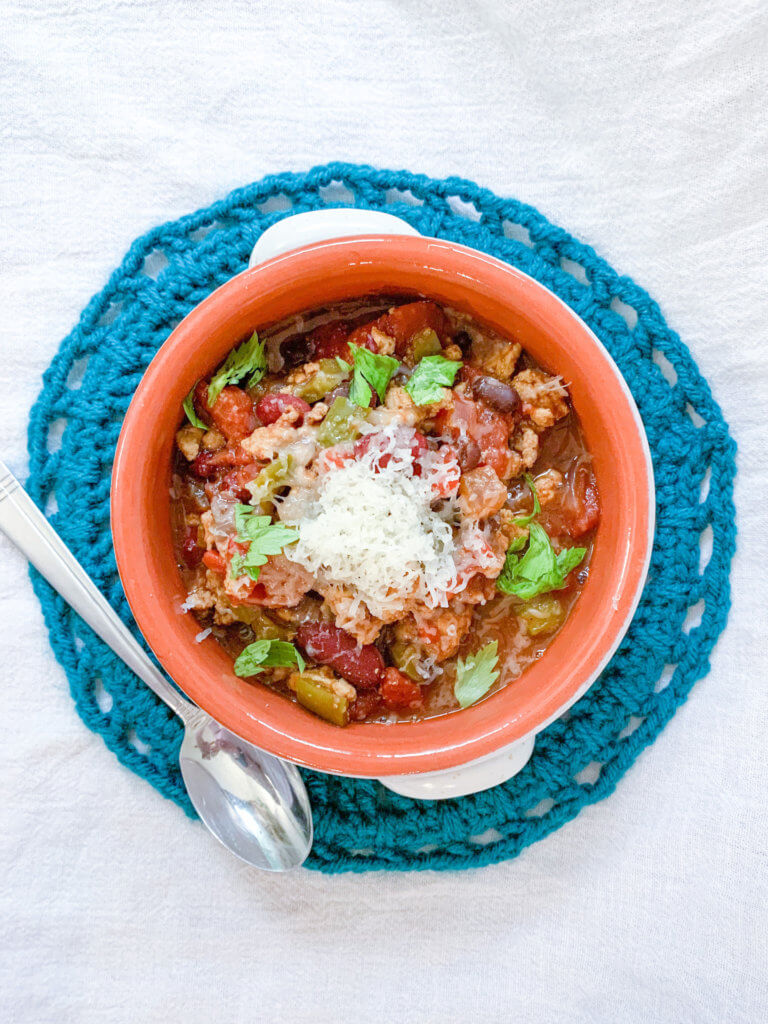 Cozy bowl of turkey chili on a teal doily topped with shredded manchego and celery greens