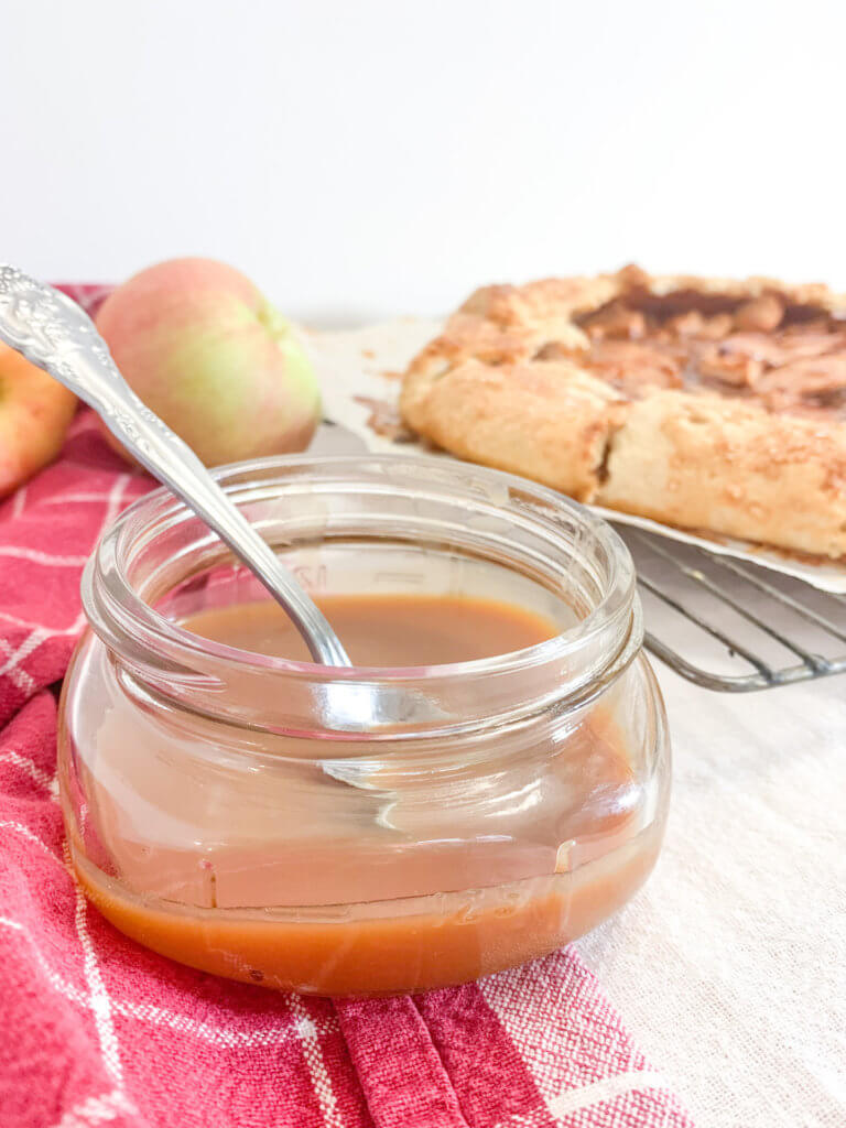 Small mason jar of caramel sauce with caramel apple galette and apples in the background