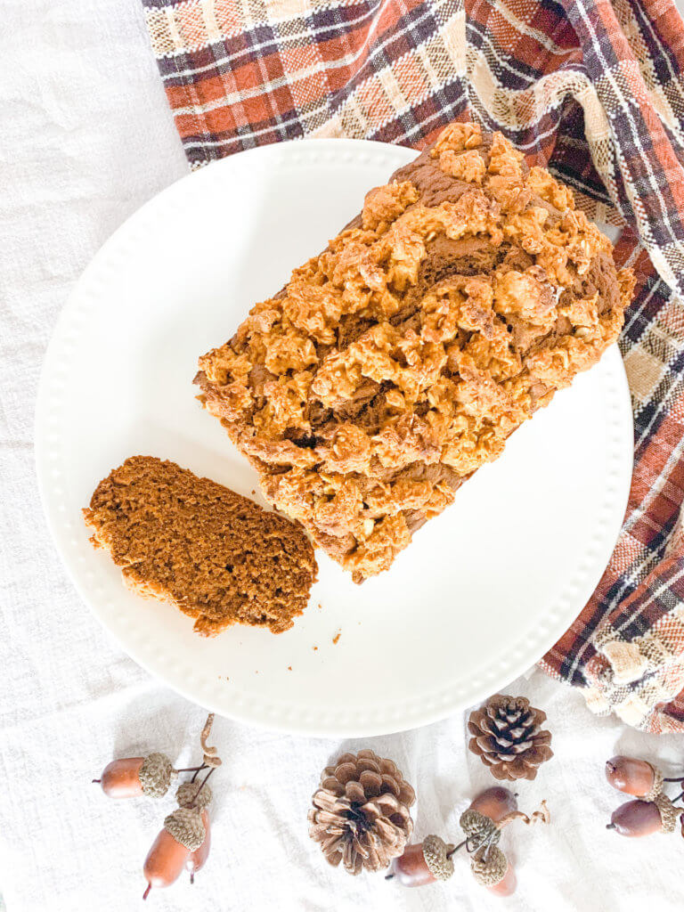 Pumpkin streusel loaf cake on a white plate with a slice cut off of it, surrounded by pinecones and acorns with a plaid dishtowel