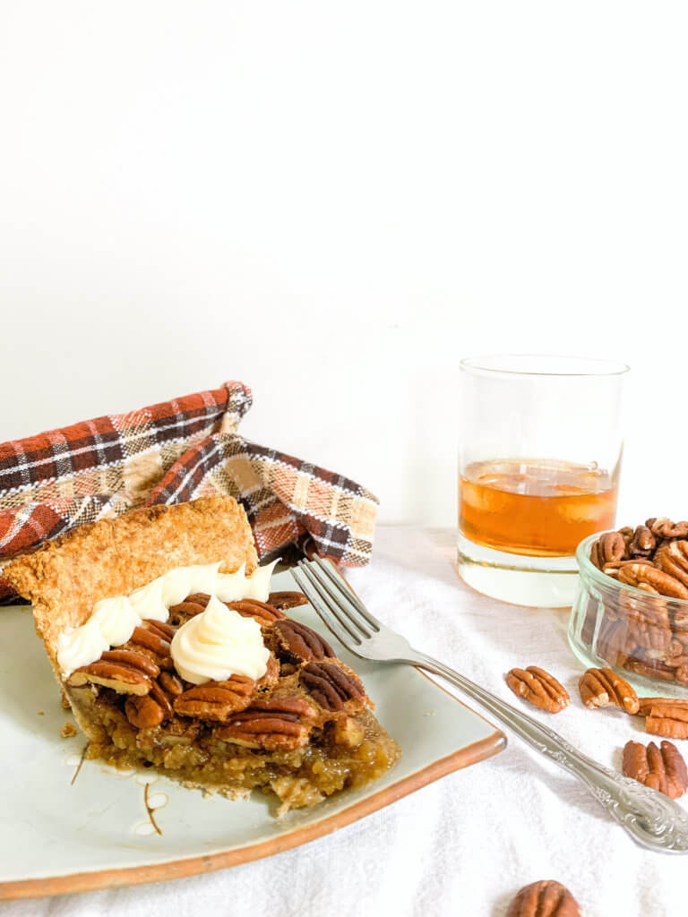 Slice of maple bourbon pecan pie with glass of bourbon and bowl of pecans