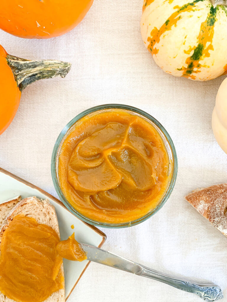 Pot of pumpkin butter beside a slathered piece of bread with some butter still on the knife.
