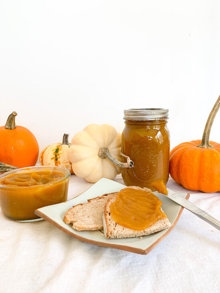 Pumpkin butter in a pot and a jar with some on a slice of toast and a bit still on the knife. Pumpkins around for decoration.