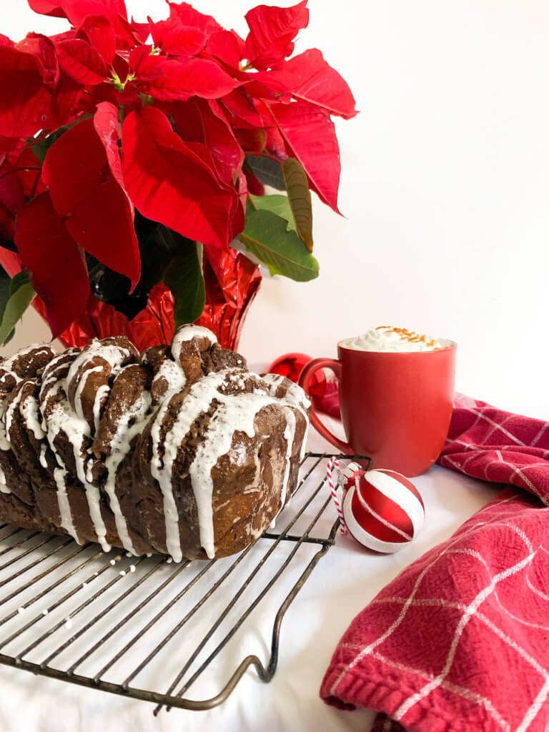 Front shot of the hot chocolate babka with marshmallow glaze under a poinsettia and next to a mug of cocoa and a red plaid dish towel.