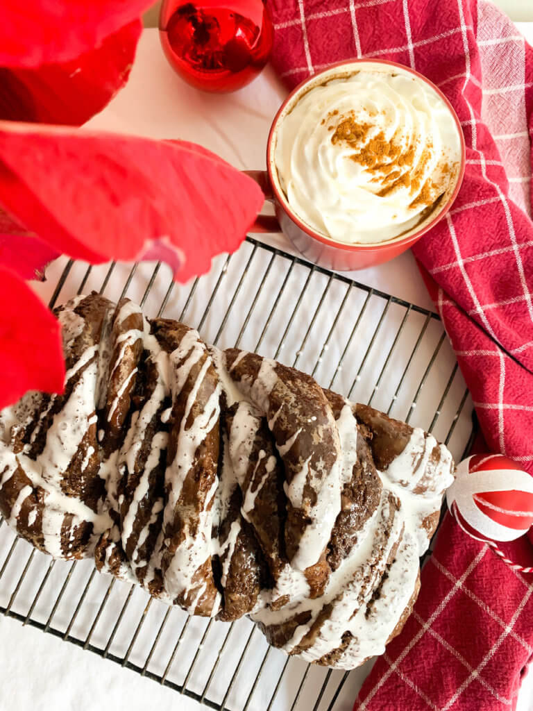 Hot cocoa babka with marshmallow glaze next to a mug of cocoa topped with whipped cream and cinnamon plus christmas decorations