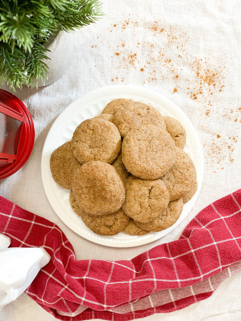 Overhead shot of vegan snickerdoodles with a sprinkle of cinnamon, a small Christmas tree, a red lantern, a white geometric bear, and a red plaid towel.