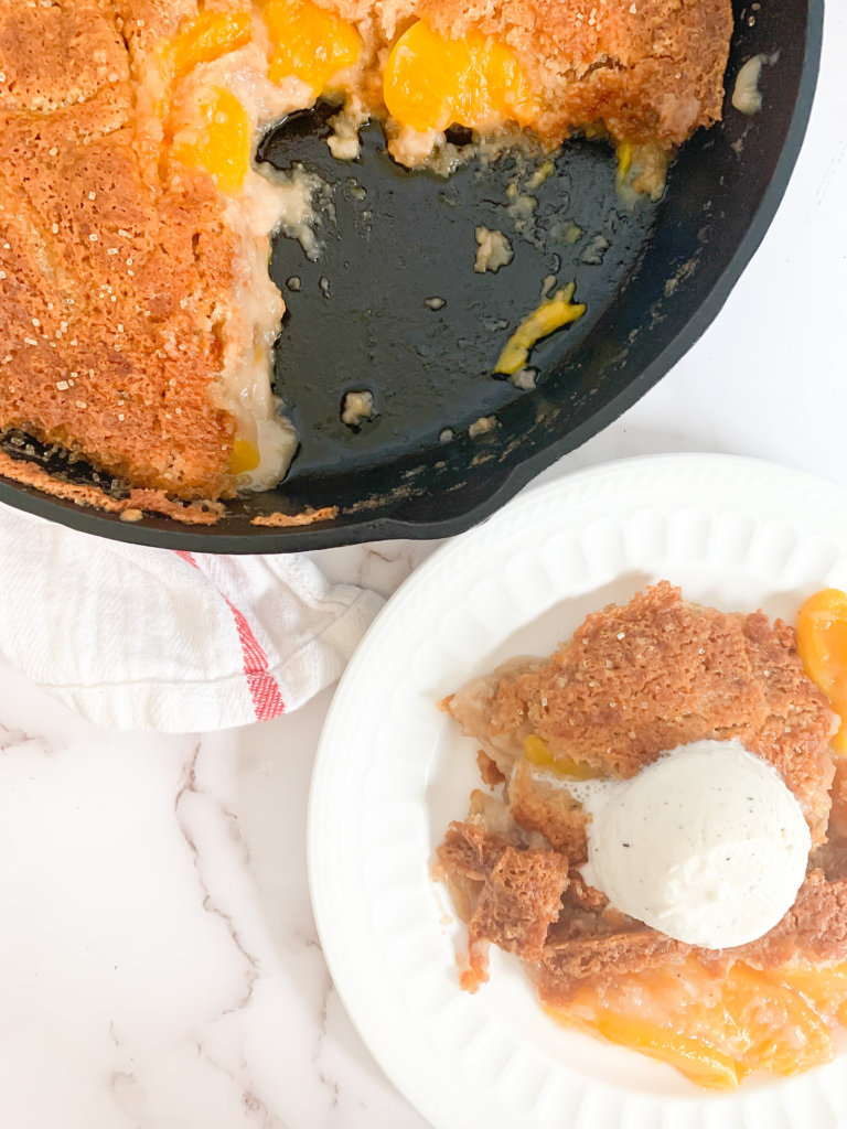 Small plate of peach cobbler with vanilla ice cream next to cast iron pan of cobbler