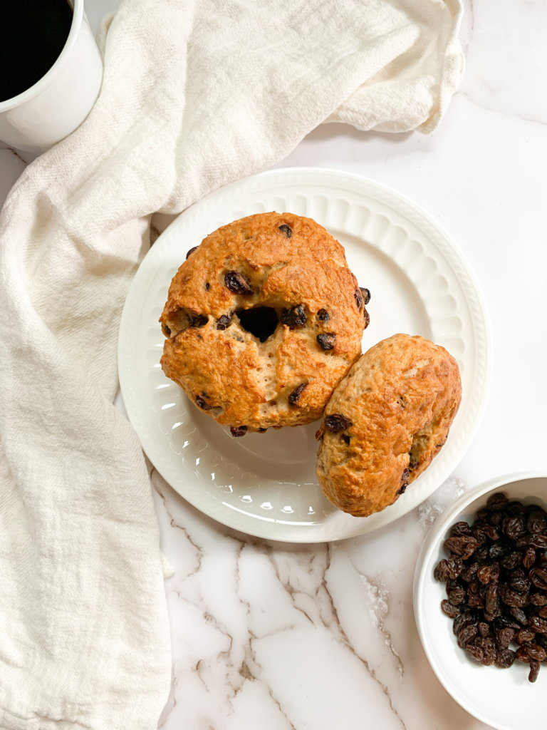 Cinnamon Raisin Bagels stacked on a plate with a cup of black coffee, a white dish towel, and a bowl of raisins on a marble counter.