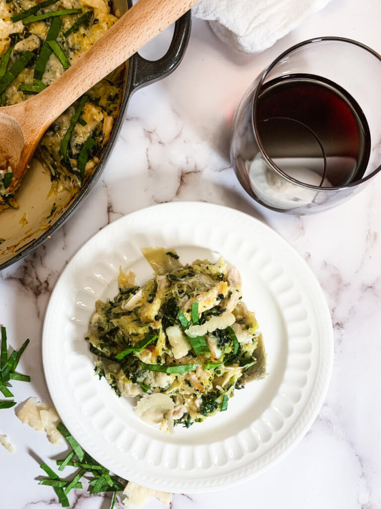 Serving of spinach artichoke casserole on a white plate with a glass of red wine