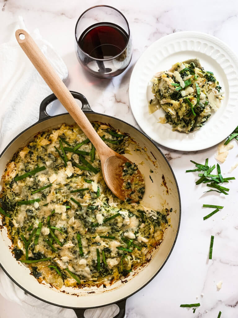 Serving of spinach artichoke casserole on a plate next to casserole dish and a glass of red wine