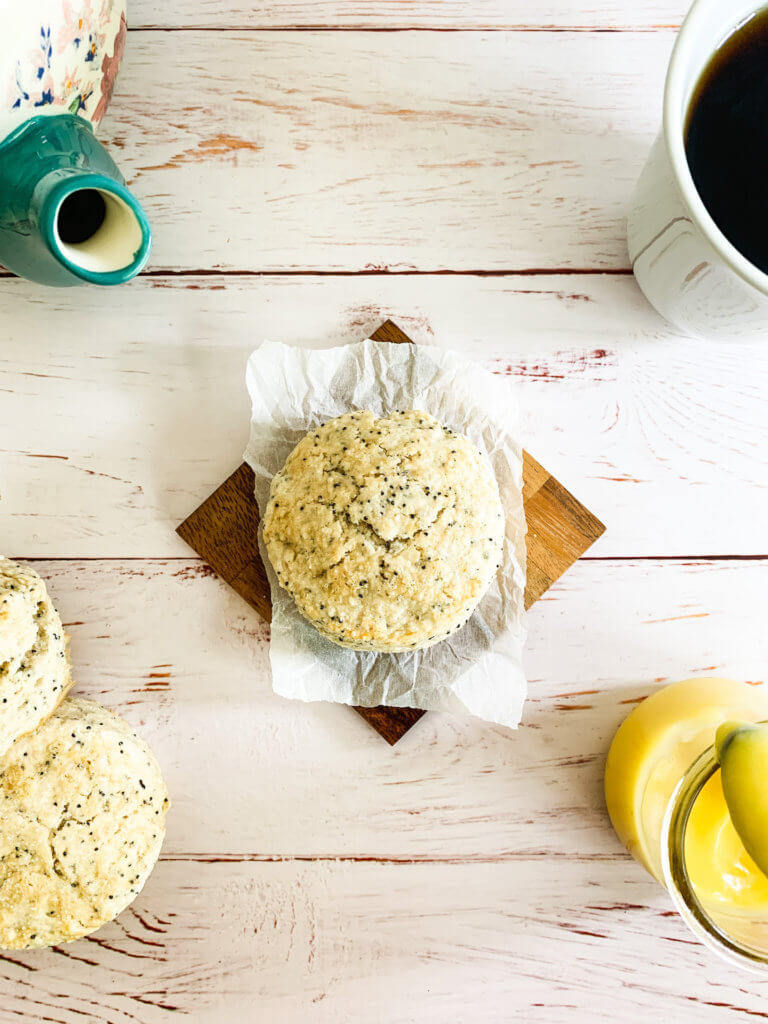 Overhead of an almond poppyseed scone in the center of a teapot, a cup of tea, a pot of lemon curd, and a pile of scones.