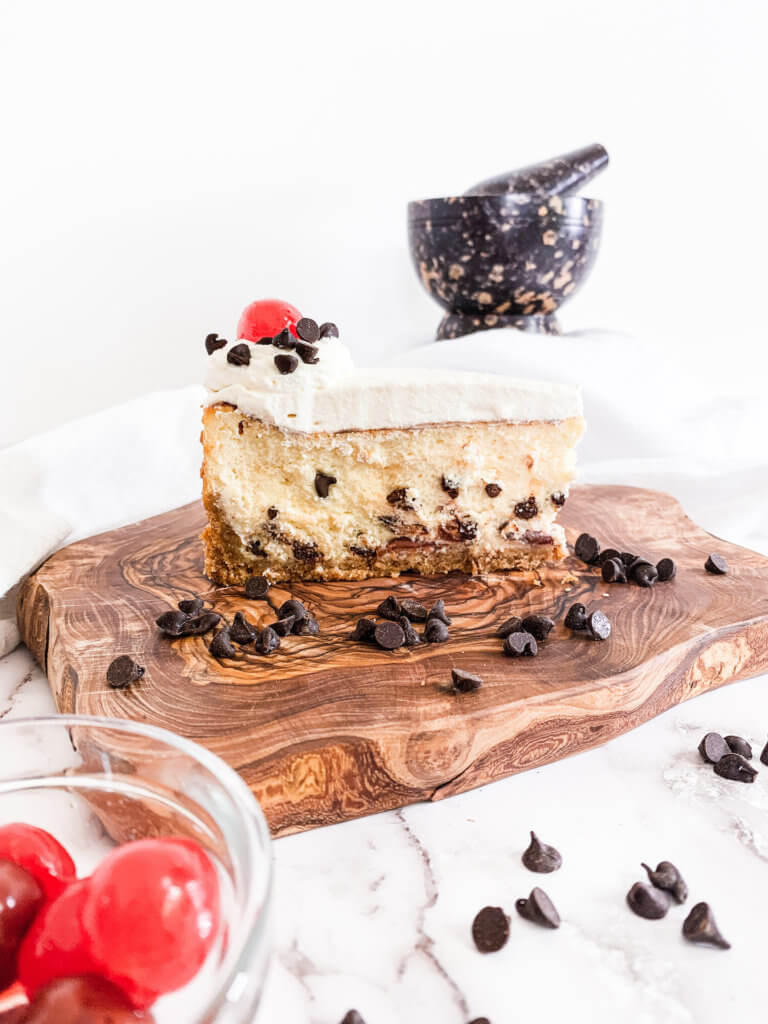 Slice of cannoli cheesecake with chocolate chips on an olive wood slab