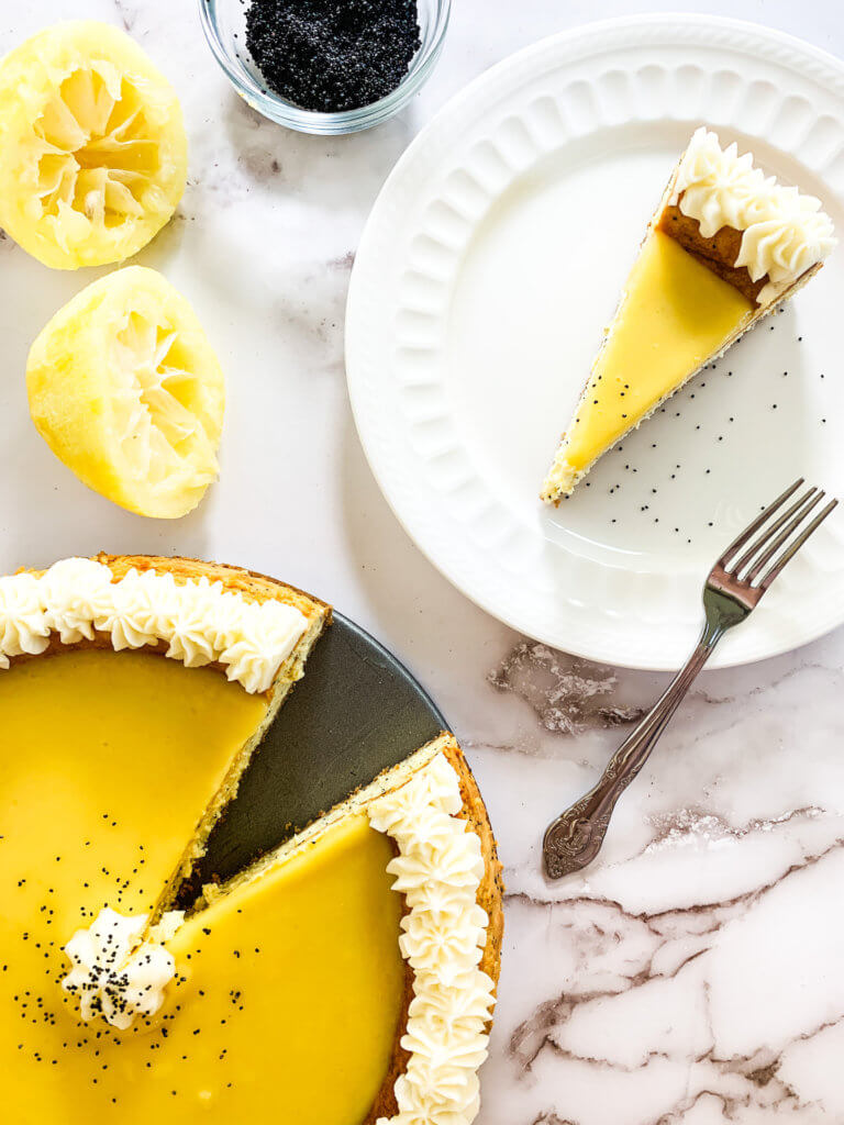 Slice of lemon poppyseed cheesecake next to the whole cake with squeezed, zested lemons and a bowl of poppyseeds
