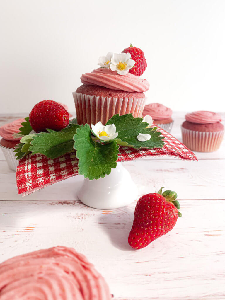 Strawberry cupcake on a pedestal with strawberry leaves, blossoms, and fresh berries