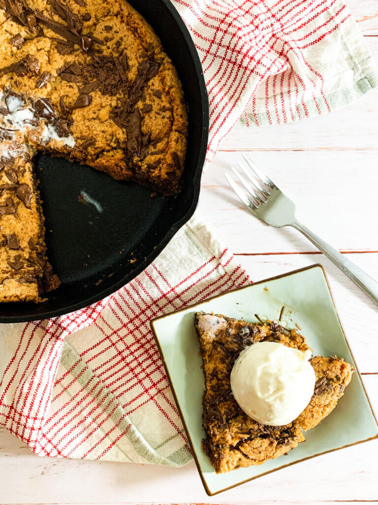 Slice of a chocolate chunk skillet cookie with vanilla ice cream on top on a light blue square plate with a red plaid napkin 