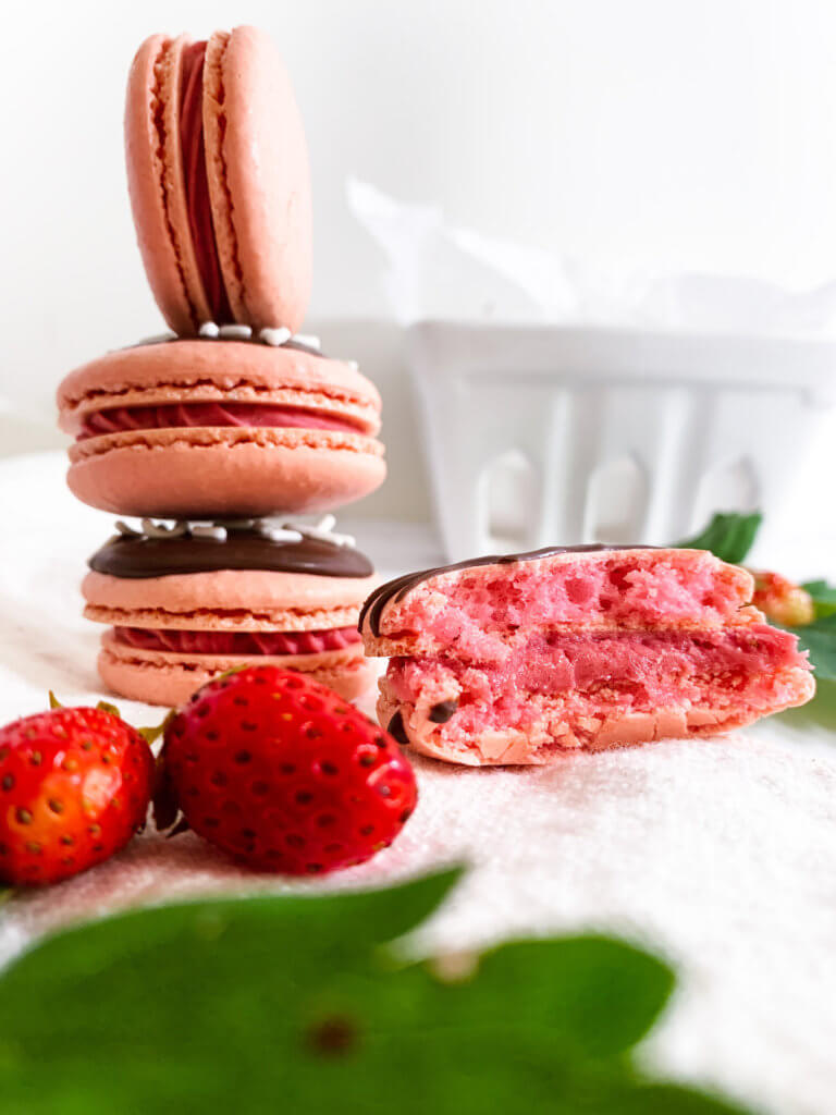 Stack of strawberry cheesecake macarons next to a half-eaten macaron with some little strawberries and strawberry leaves