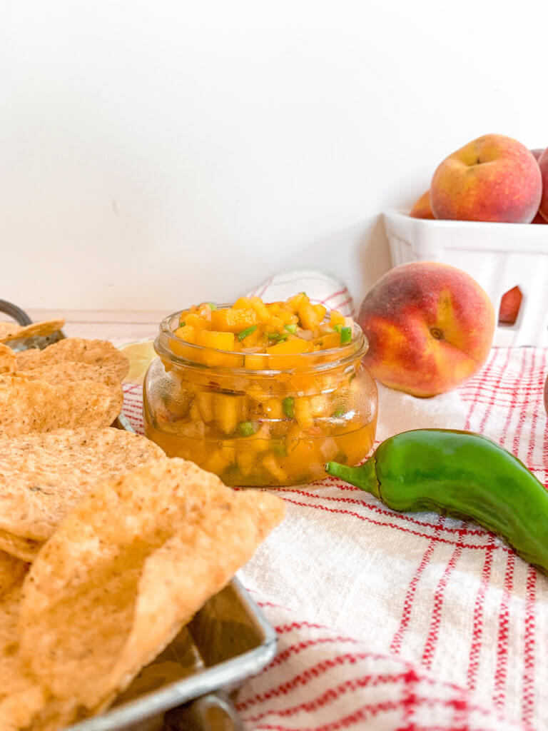 front view of spicy peach salsa in an 8 ounce short jar with tortilla chips, jalapeños, and peaches
