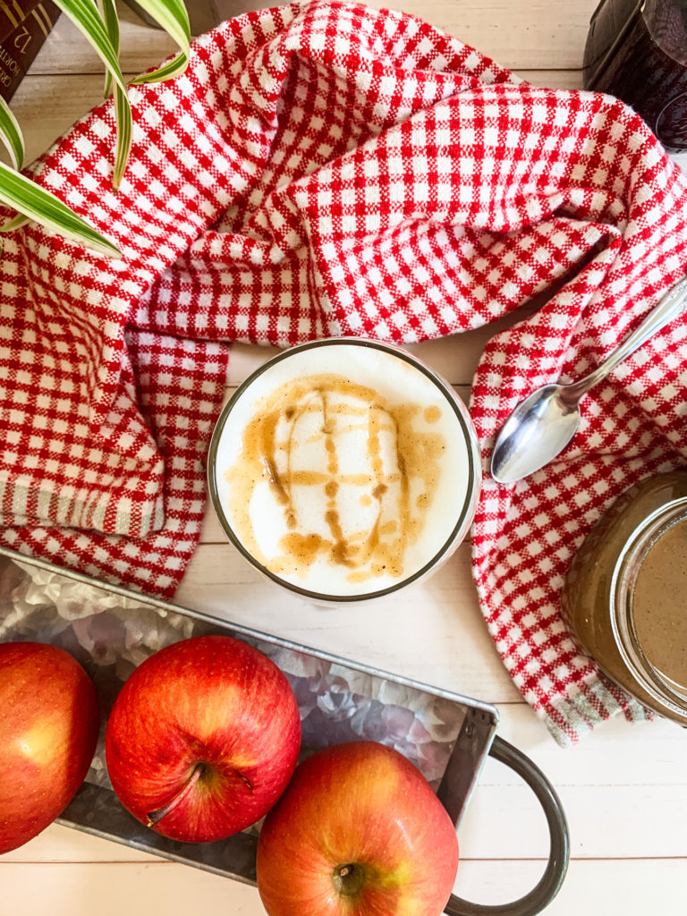 Apple crisp macchiato with apple spice caramel drizzle on top surrounded by a red check dish towel and honey crisp apples