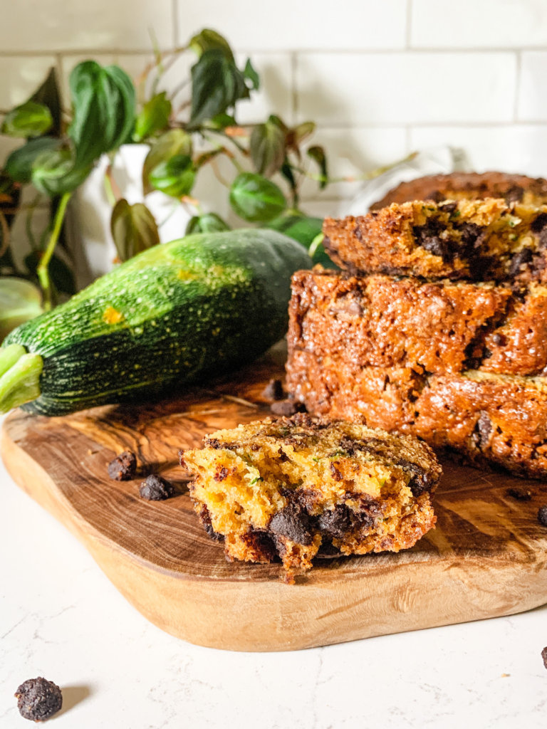 close up of a bite-sized piece of chocolate chip zucchini bread in front of a stack of slices of zucchini bread with a zucchini on an olive wood cutting board