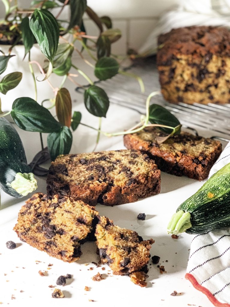 Torn slice of chocolate chip zucchini bread with crumbs and chocolate chips surrounding with zucchini and ivy around