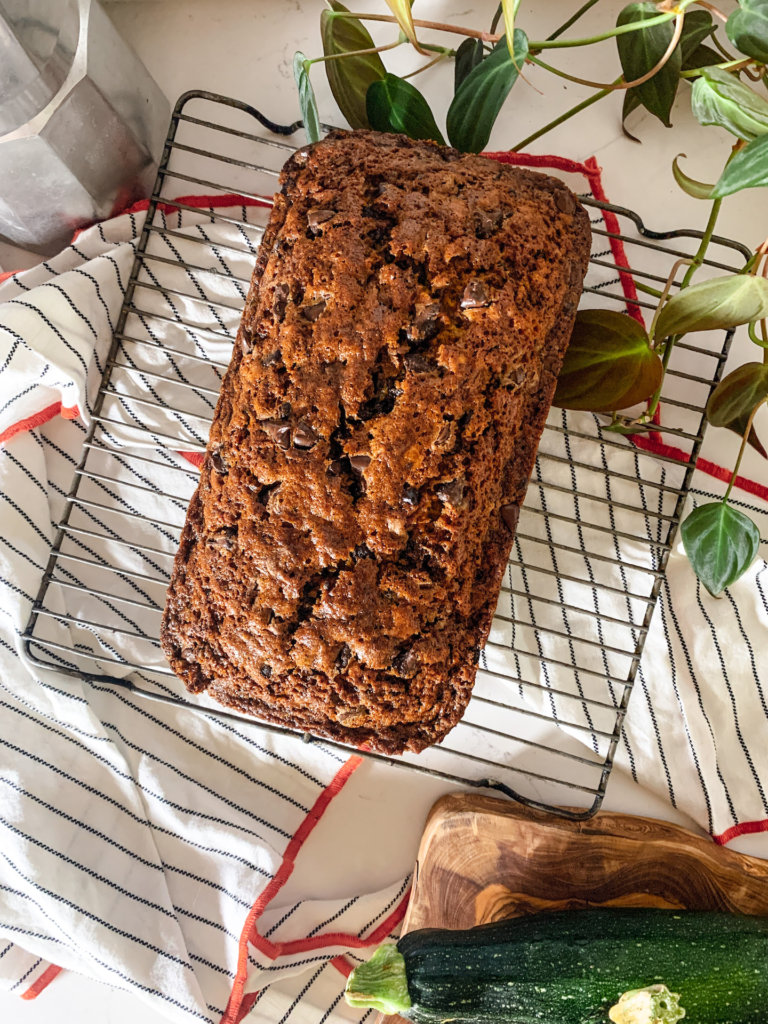 Loaf of chocolate chip zucchini bread on a cooling rack with pothos ivy and zucchini