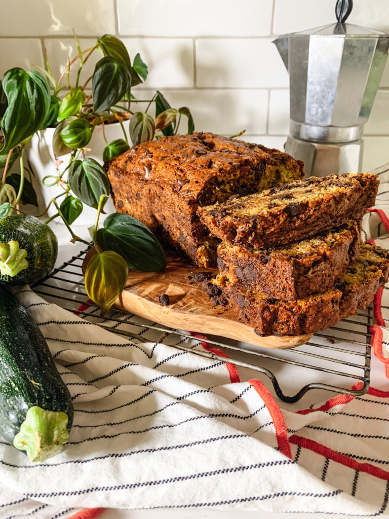 Stack of sliced chocolate chip zucchini bread next to half loaf with an espresso pot in the background and two zucchini in the foreground