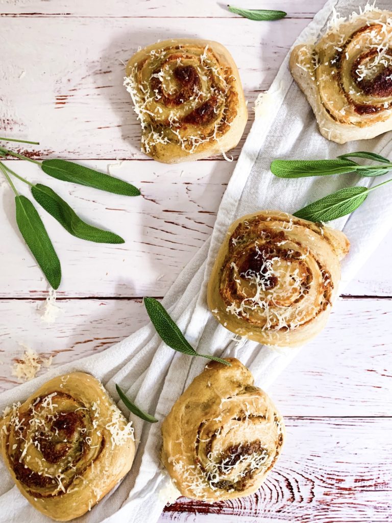 Asiago pesto buns on a white cloth on a white-washed wood surface with sage leaves
