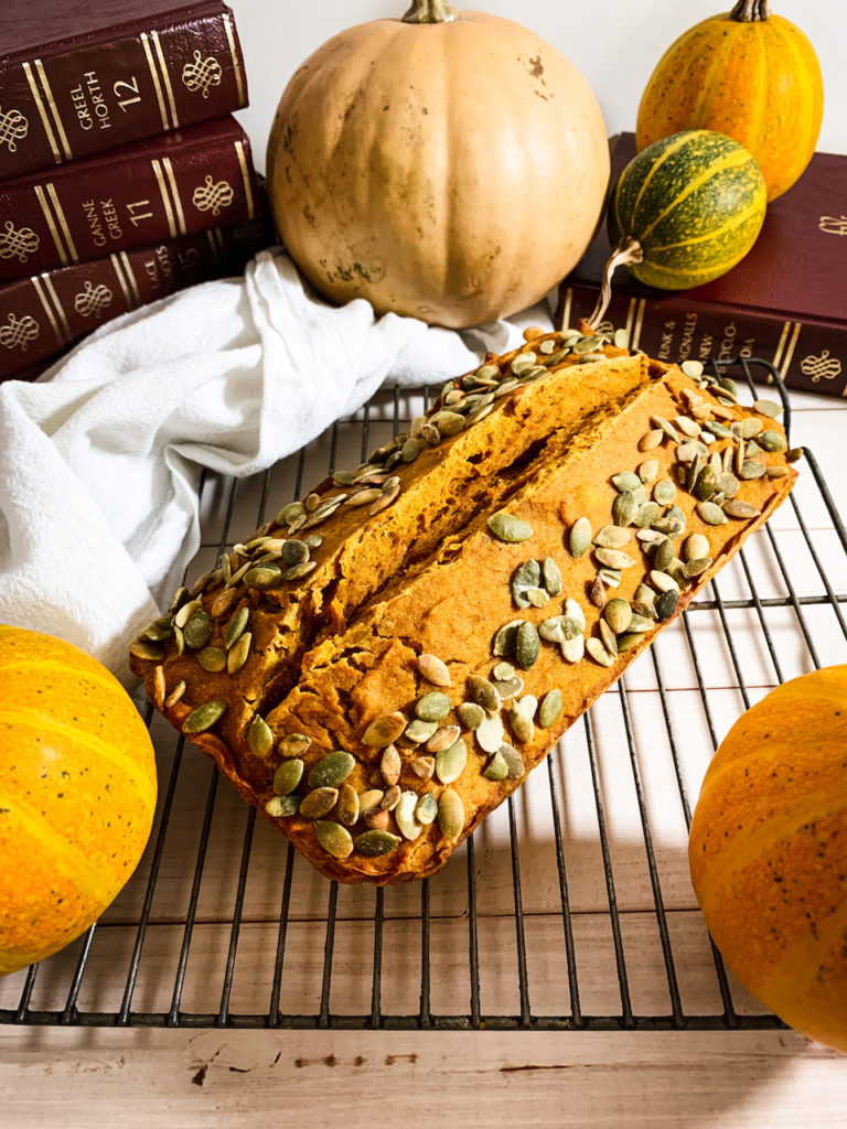 Loaf of maple pumpkin bread with pepitas on top on a cooling rack surrounded by baby pumpkins and leather-bound encyclopedias